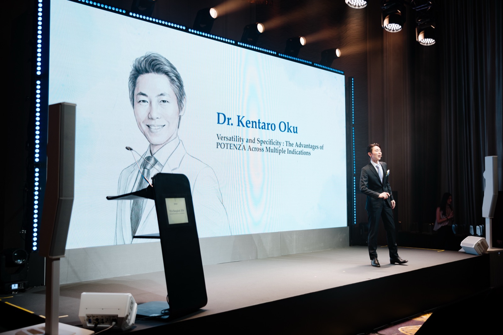 TEA Bangkok 2026の演題スライドとステージに立つヒルズグレイスクリニック院長 奥謙太郎